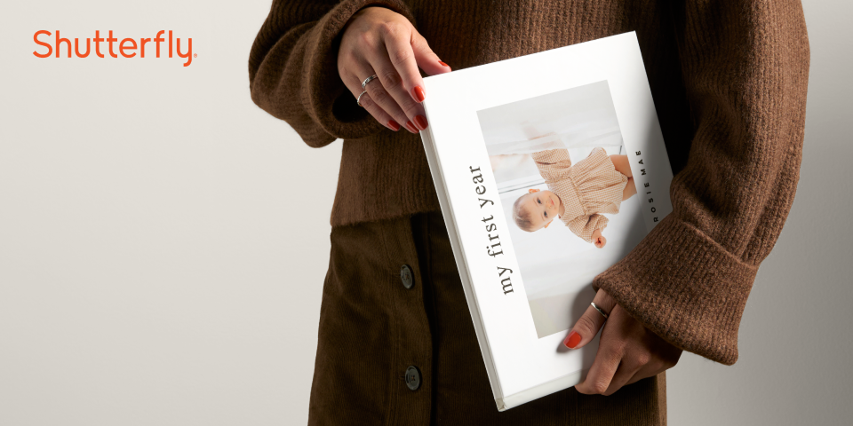 Close-up image of a person holding a beautiful Shutterfly photo book. The book is title, “My First Year” and has an image of a baby on the cover. To the left side of the frame, there’s a Shutterfly logo.