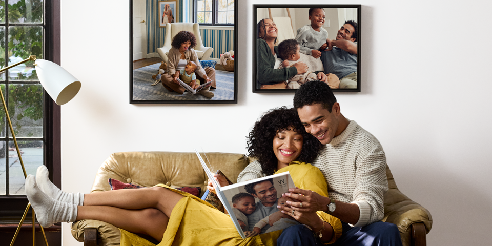 A happy couple smiles broadly as they flip through a large Shutterfly photobook. On the wall behind them, there are beautifully printed photos.