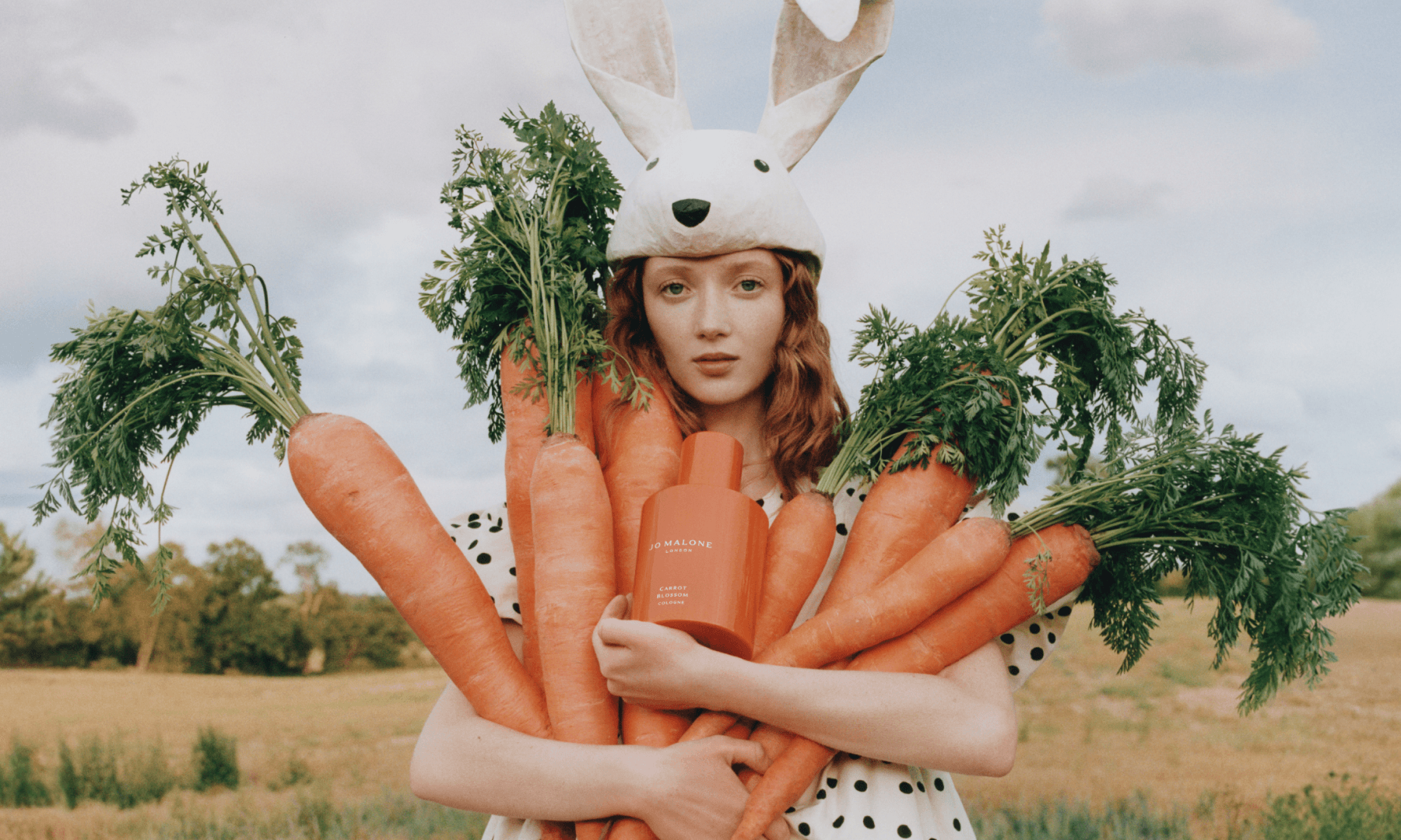 Woman holding a few large carrots and a bottle of Jo Malone Carrot Blossom Cologne, wearing a rabbit hat and polka-dot clothing in the countryside.