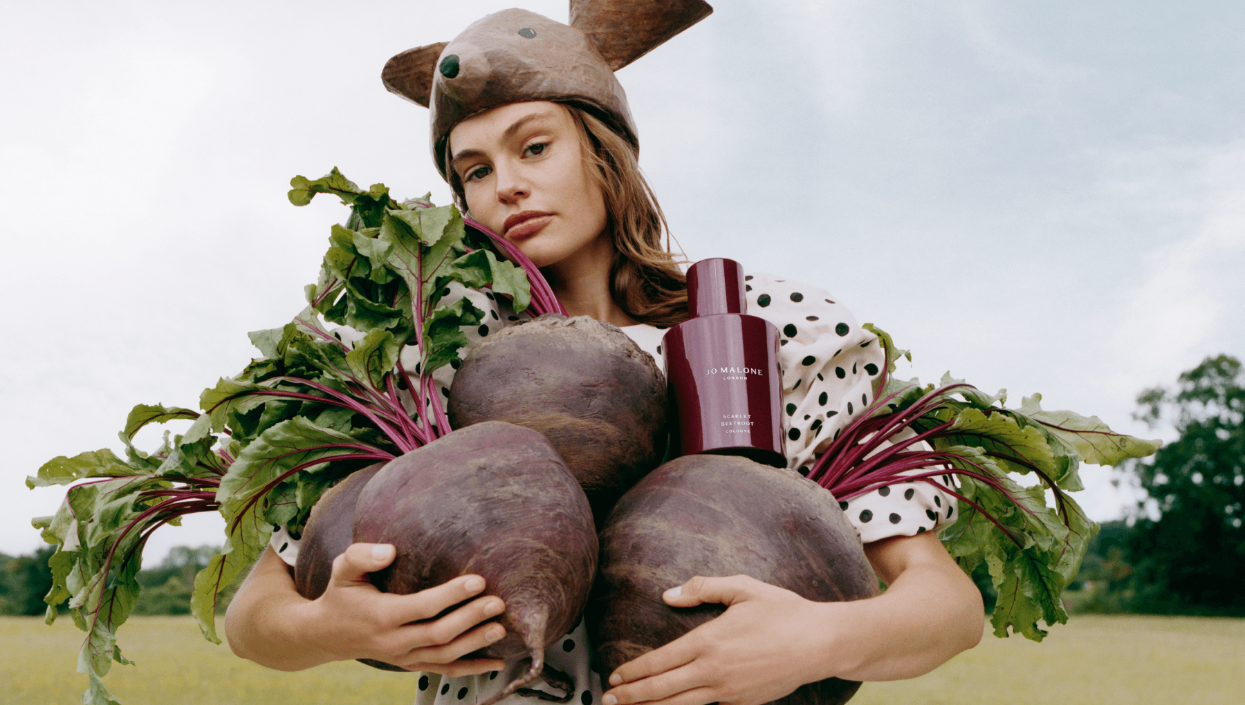 Woman holding four giant beetroots and a bottle of Jo Malone Scarlet Beetroot Cologne, wearing an animal hat and polka-dot clothing in the countryside.