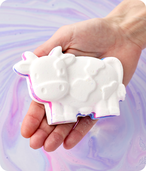 image: Toby’s Magic Cow – A hand holding a cow shaped bath bomb in front of colorful bath art water